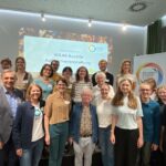 etwa 30 Menschen stehen lächelnd vor einer Leinwand auf der steht WEAll Austria Auftaktevent