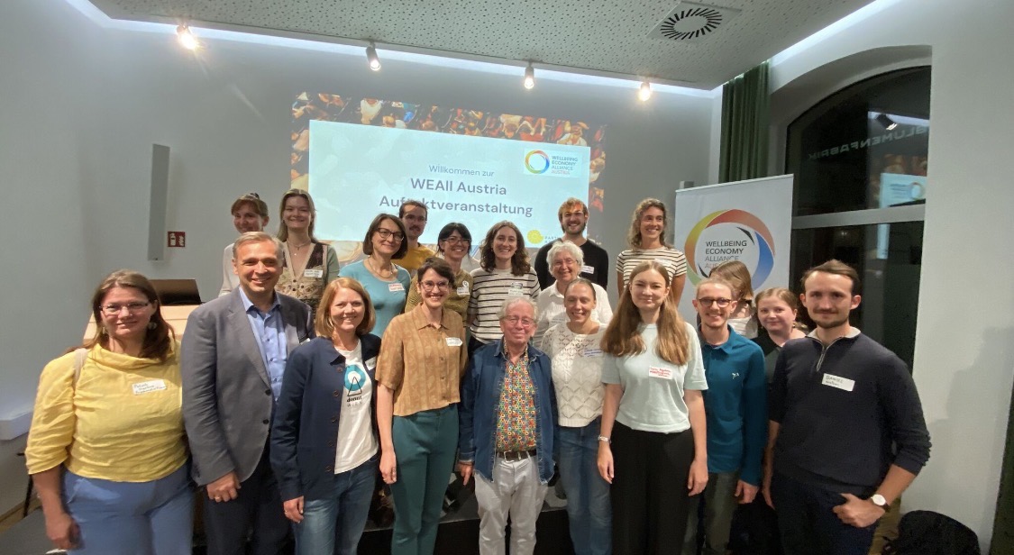 etwa 30 Menschen stehen lächelnd vor einer Leinwand auf der steht WEAll Austria Auftaktevent
